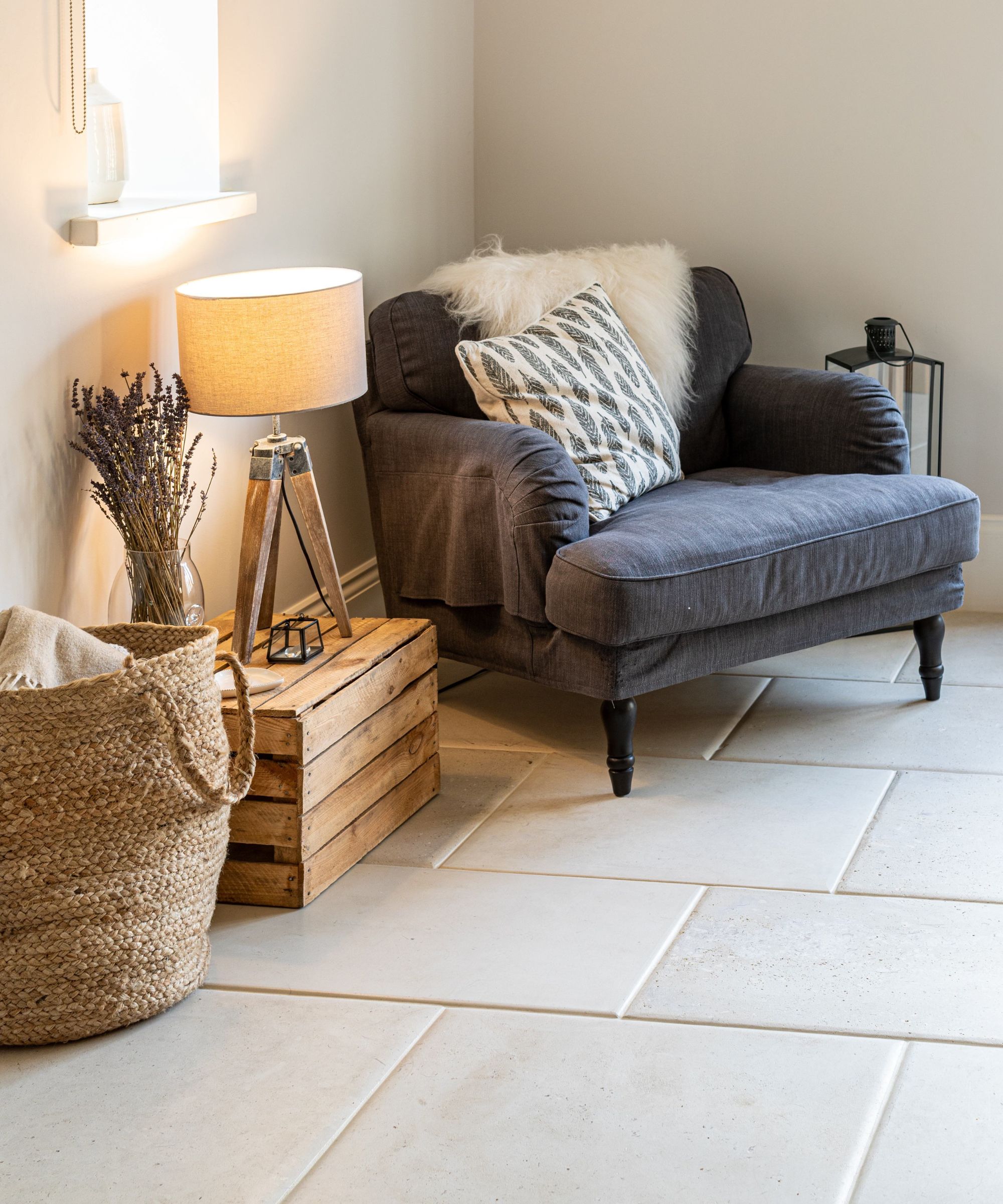 A cosy living room corner featuring a dark grey armchair with patterned cushions, a wooden crate used as a side table with a tripod lamp, a woven basket and large pale stone floor tiles.