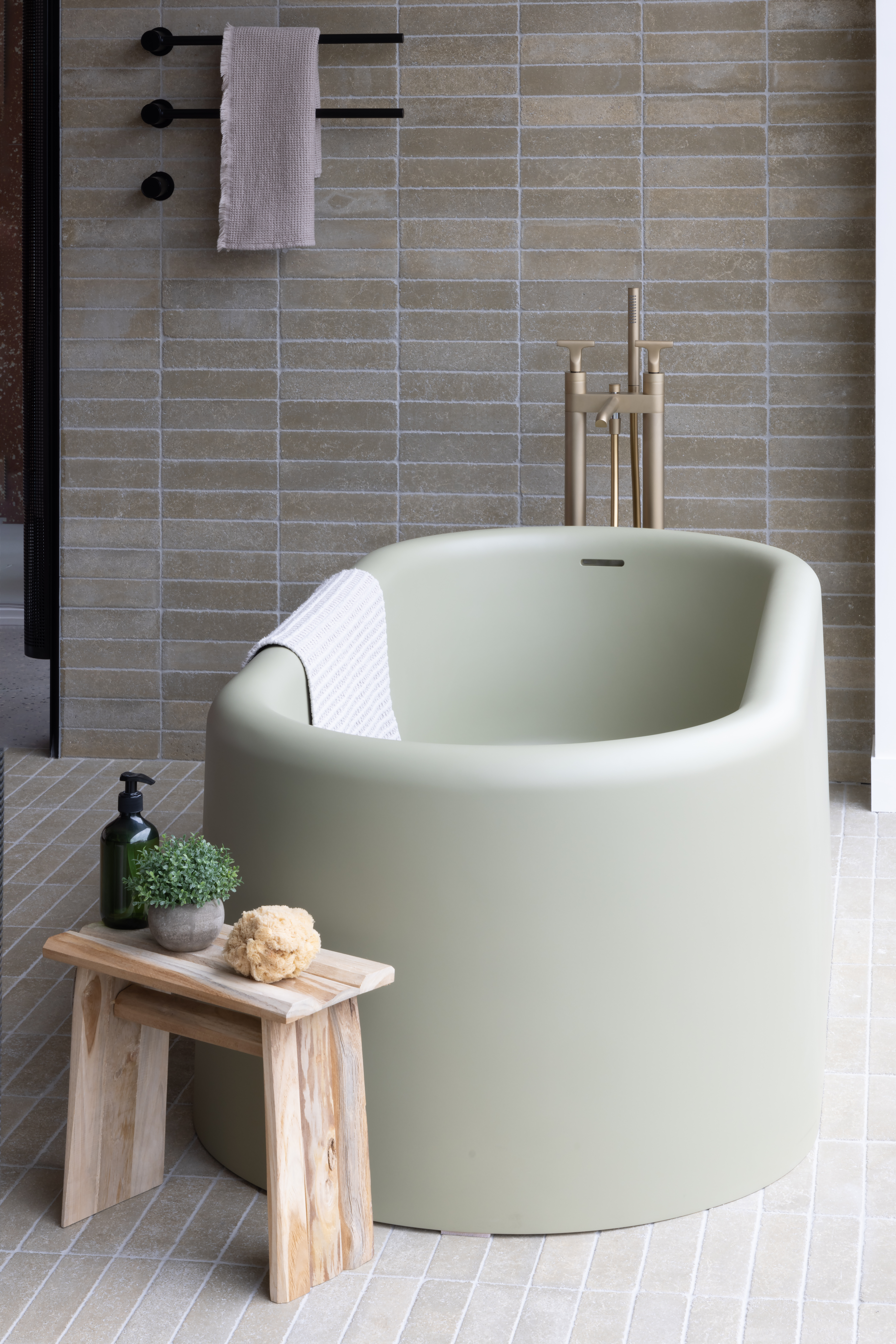 Pale green stand alone tub with copper brassware in a stone tiled bathroom. In front of the tub stands a small wooden table