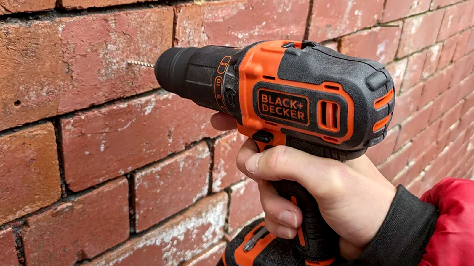 Person holding orange and black Black &amp; Decker drill drilling into red brick wall 