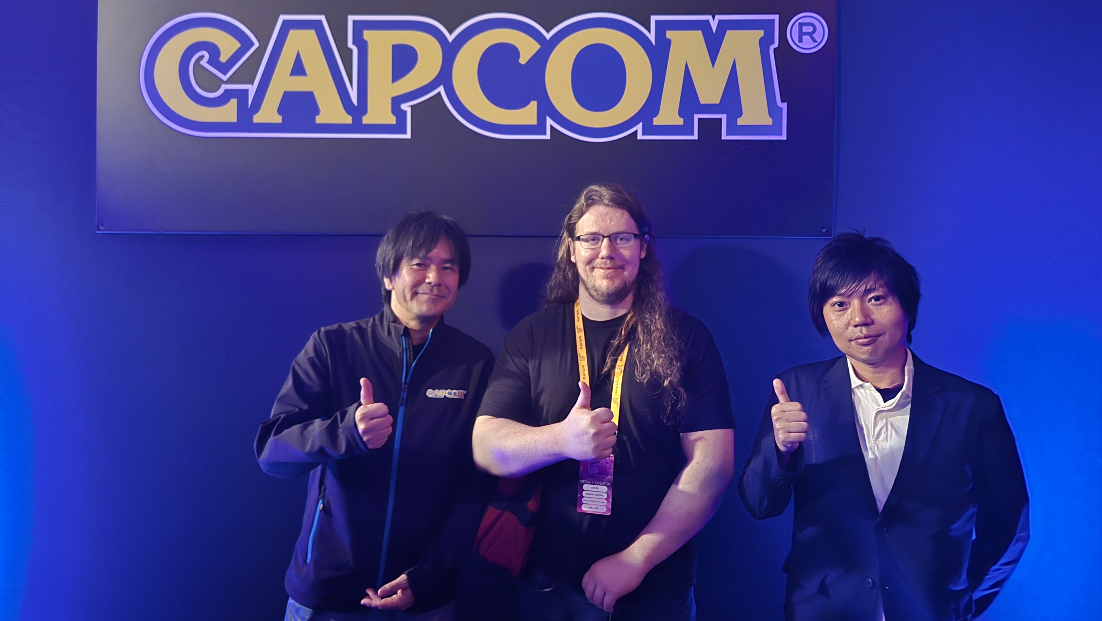 Three men standing up giving a "thumbs-up" hand gesture under a sign that says "Capcom"