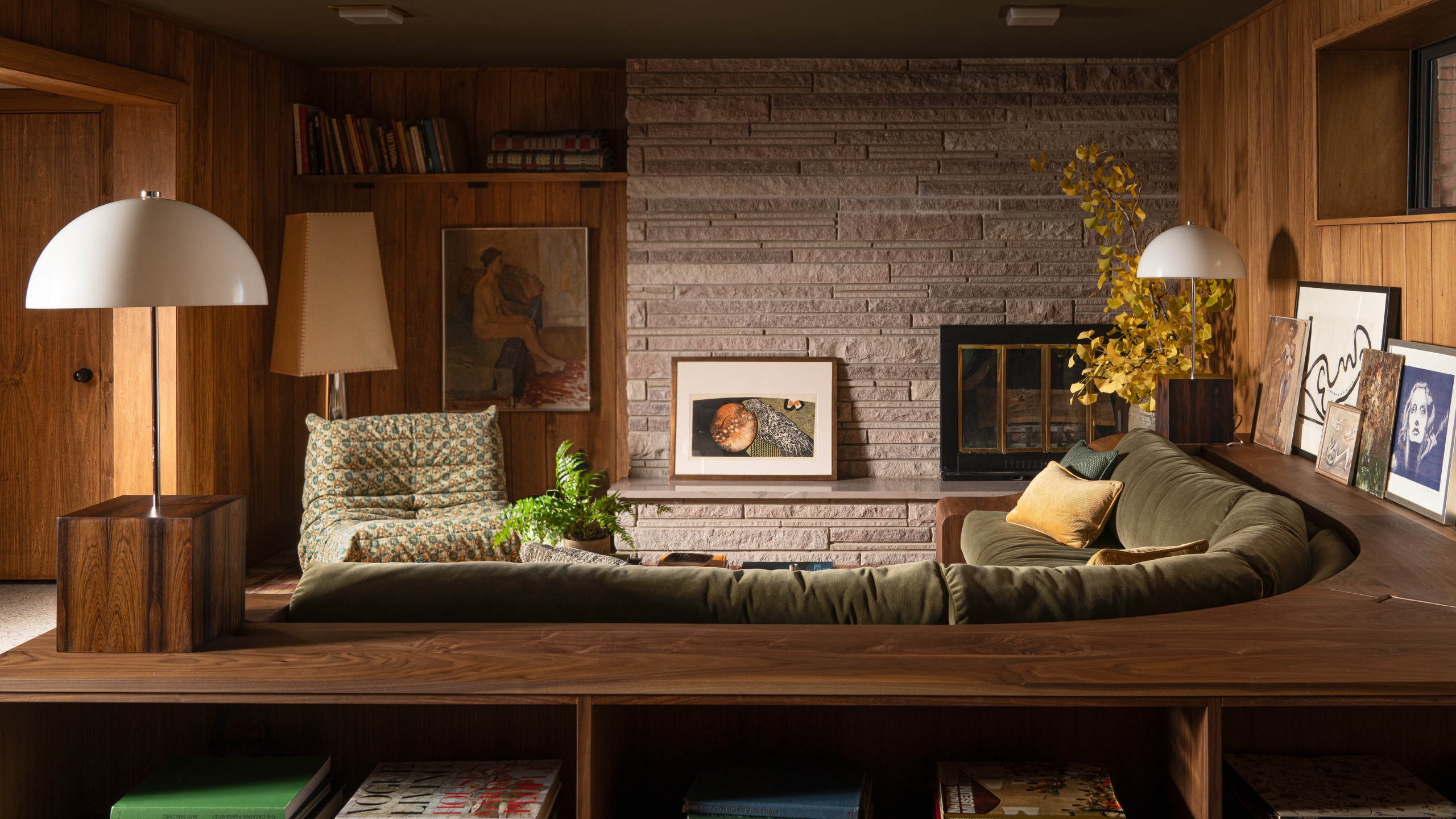 A mid-century family room with wood paneled walls, a stone fireplace, and a built-in sunken corner sofa with olive green upholstery