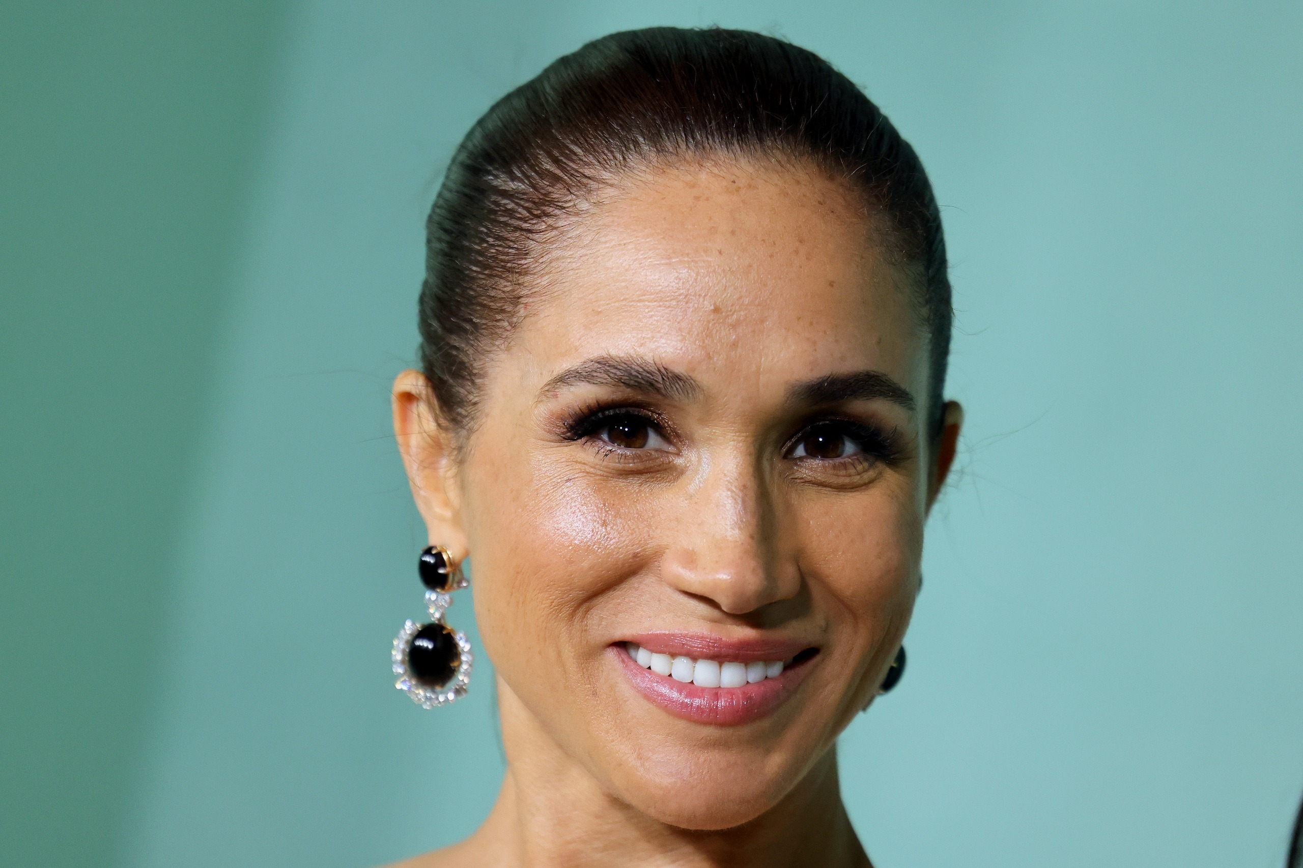 A headshot of Meghan Markle wearing dangling black earrings with her hair slicked back