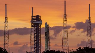 a white rocket stands at a launchpad surrounded by four tall lightning towers against an orange sky.