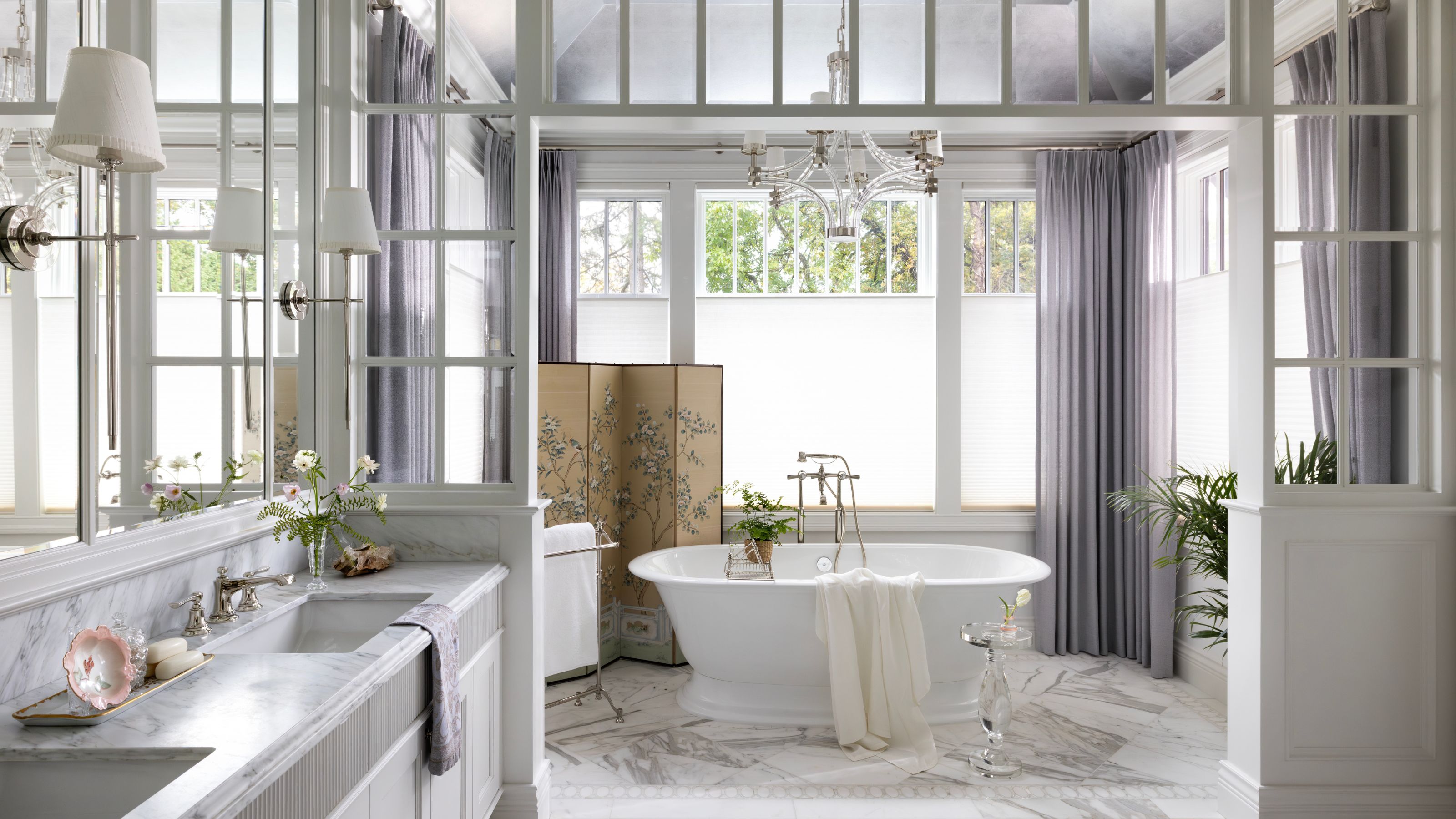 Bright luxury bathroom with a white freestanding tub, marble floors, lavender curtains, and a decorative folding screen under a glass-paned ceiling.