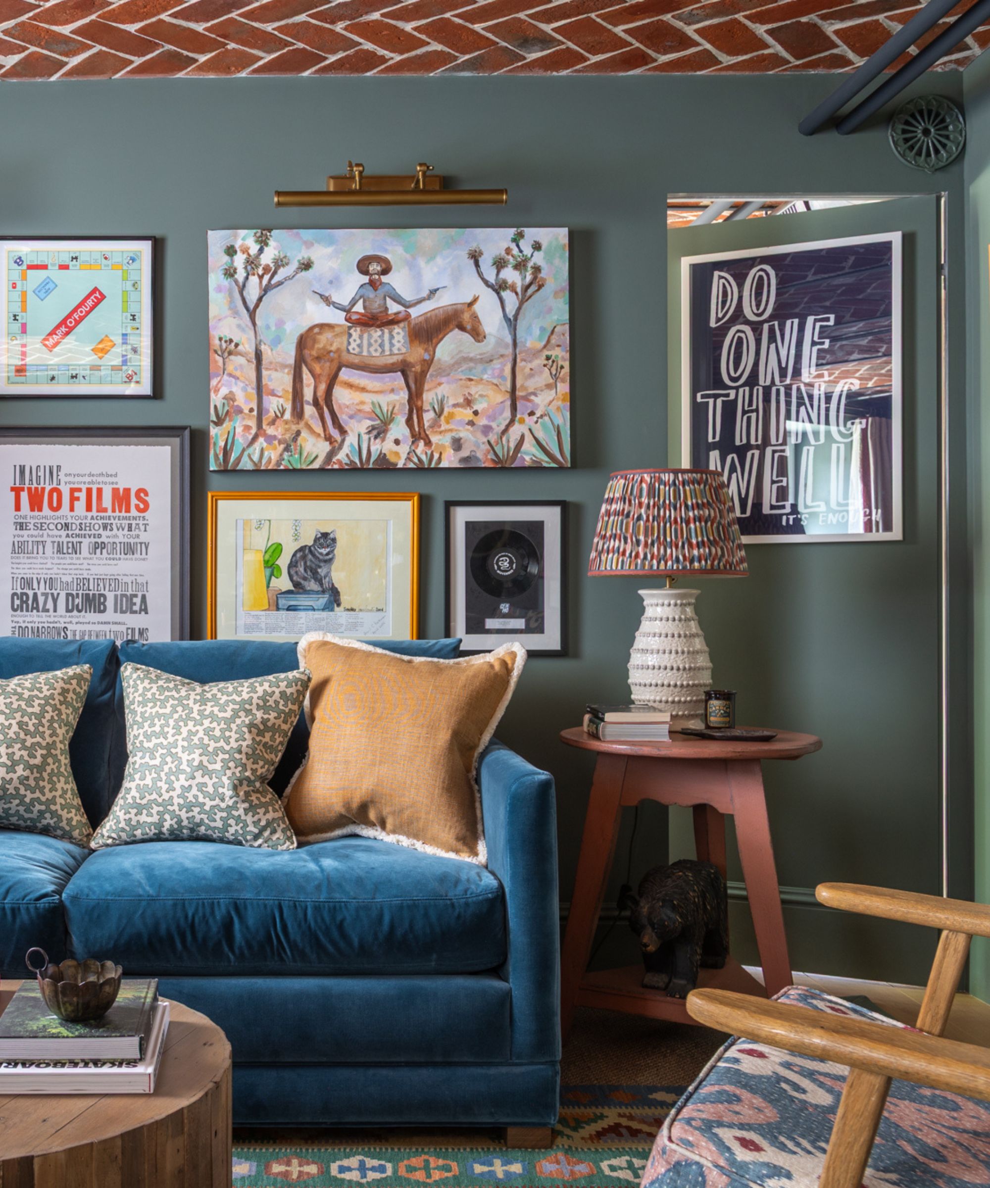 A dark teal living room with a wall covered in an eclectic gallery of mixed and matched artwork, a blue velvet sofa and coral colored round side table with a ceramic lamp.