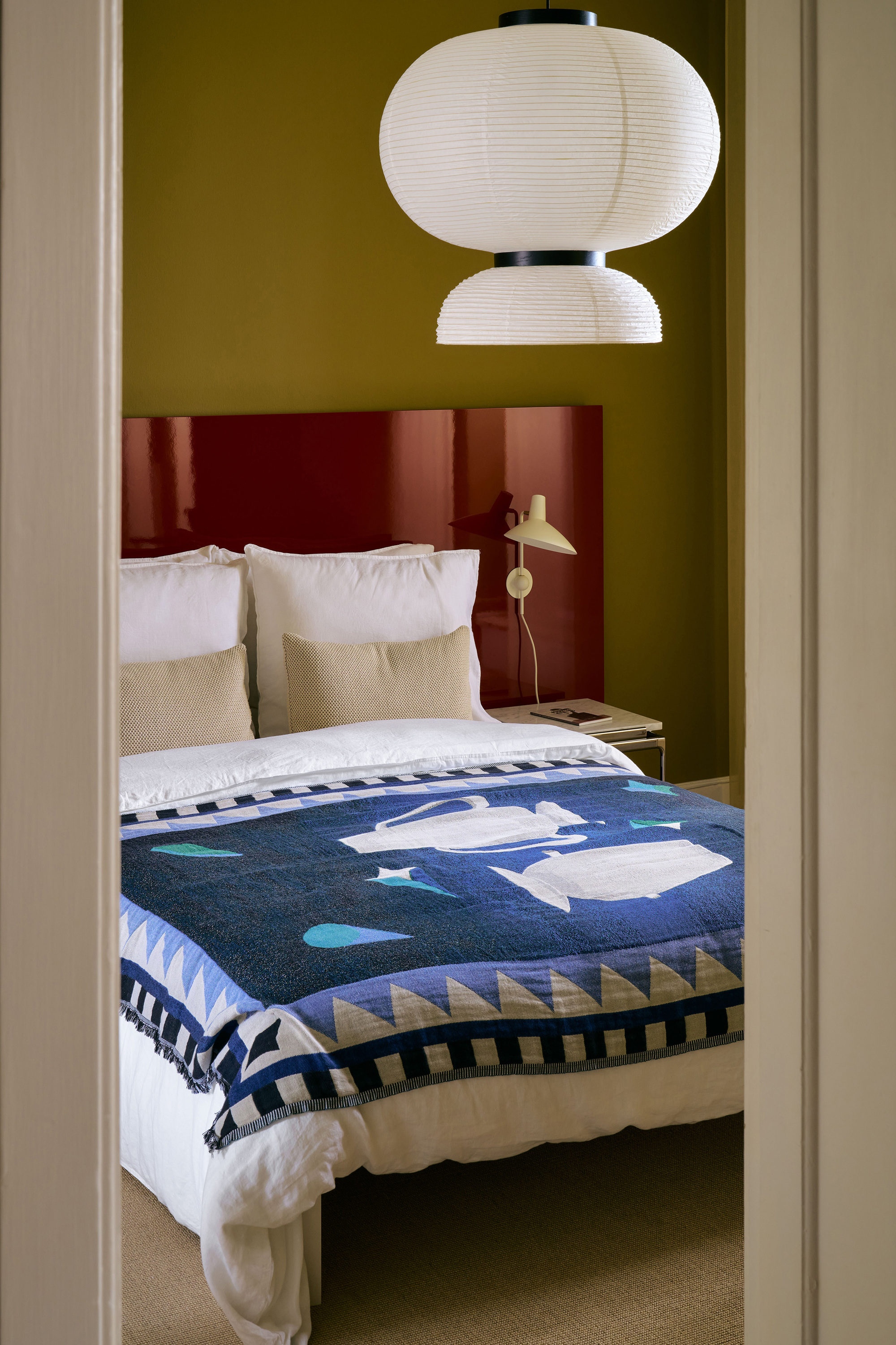 modern bedroom with green walls, red lacquered headboard, rice paper pendant light, white and beige pillows, white sheets and a patchwork quilt
