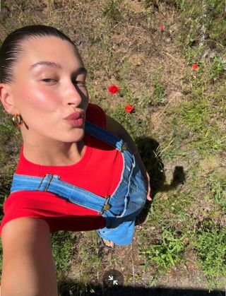 Hailey Bieber wearing a red T-shirt and baggy denim overalls in a high-up selfie in the grass.