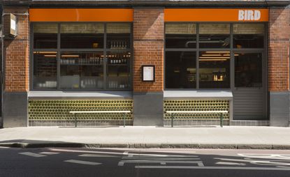 Street view of the Bird Restaurant with an orange banner above two large windows separated by brick pillars with two benches on the pavement.