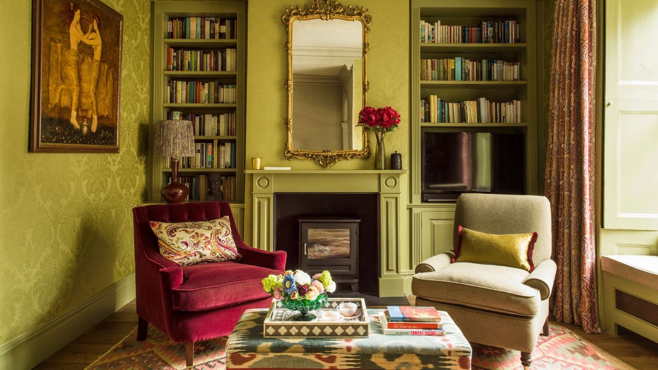 A lime green color drenched living room with contrasting patterned carpet and footstool, a burgundy velvet armchair with a matching beige upholstered one, a gold statement mirror, roses in a vase, and symmetrical color drenched bookshelves