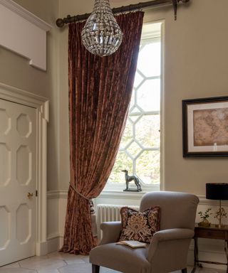 Large window dressed with brown velvet drape, white painted walls, brown armchair, stone flooring