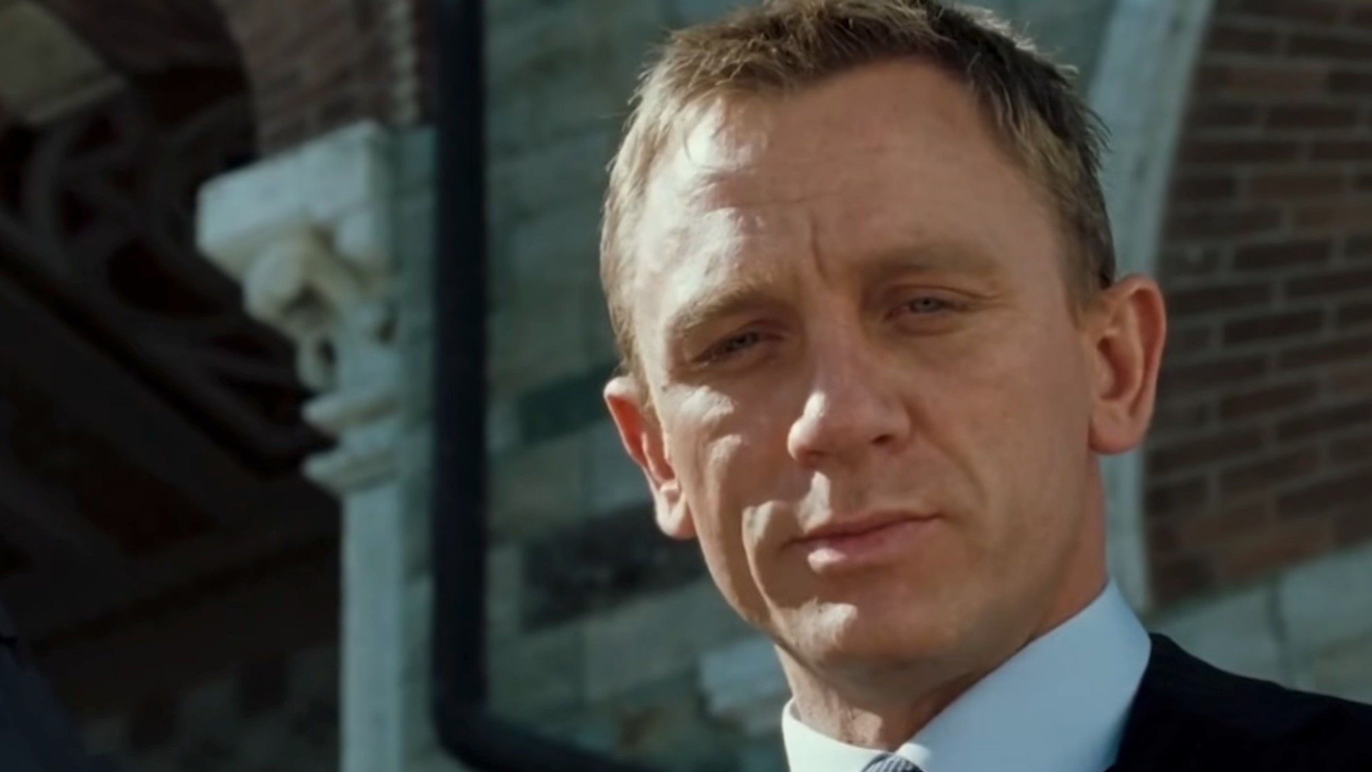 A close up of Daniel Craig as James Bond in the final scene of Casino Royale, wearing a white-collared shirt and blue suit. 
