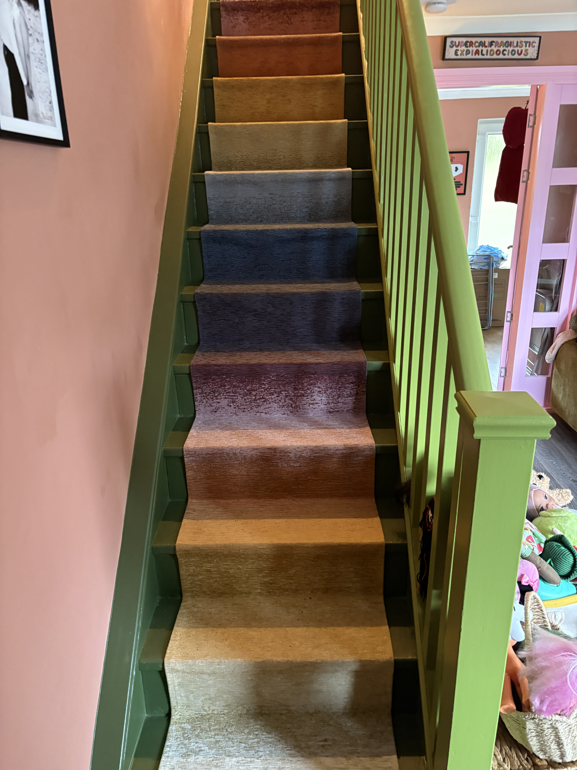 A staircase with a green painted balustrade, pink walls, and a rainbow ombre rug