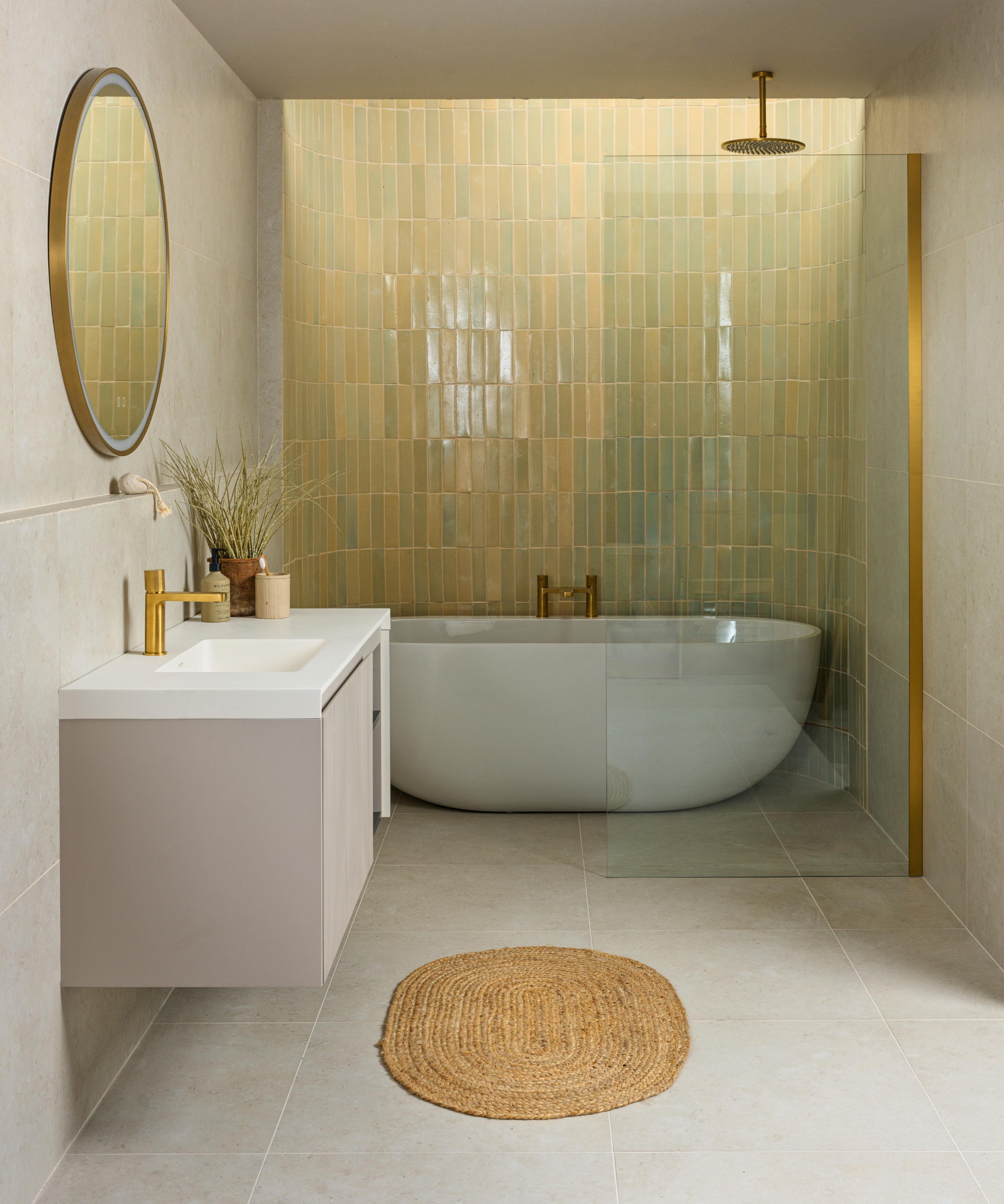Modern spa-style bathroom featuring a curved soaking tub, soft green glazed tiles, brass fixtures, pale stone flooring, and clean minimalist lines.