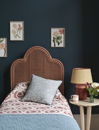 dark blue guest room bedroom with rattan headboard, artwork, Scandi style side table, table lamp, patterned bedding, blue cushion and blanket