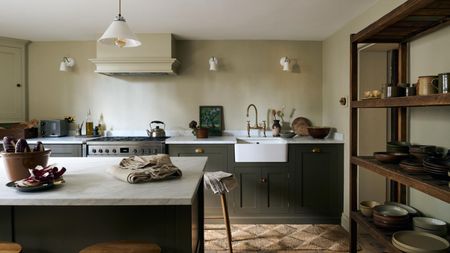 An olive green kitchen with olive green cabinets and a large kitchen island with a marble worktop. On the right side of the kitchen is an open shelf with places and mugs on top.