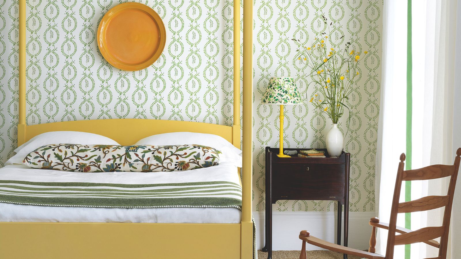 A green and yellow bedroom with white and green wallpapered walls and yellow four-poster bed, with a wooden bedside table, wooden rocking chair, large window and sheer white curtains.
