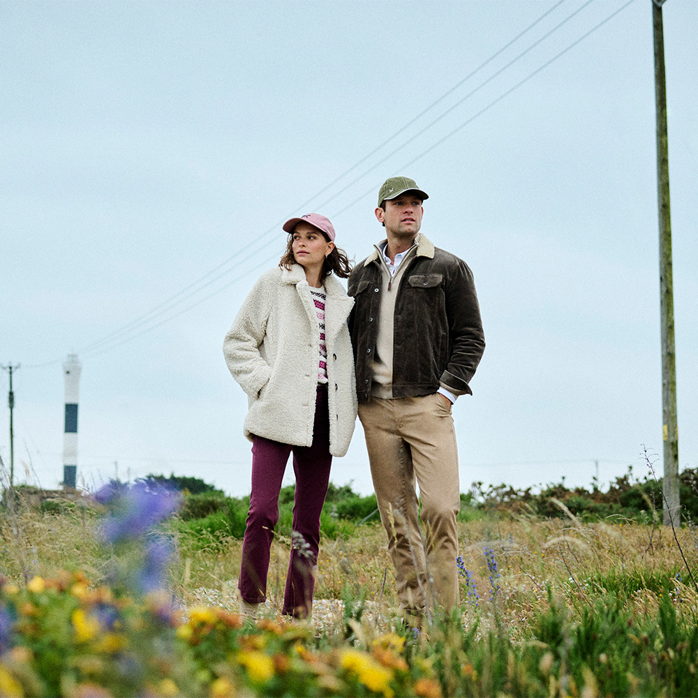 Two models in a wildflower field wearing Crew Clothing outerwear and caps