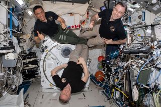 three people in dark shirts float aboard the international space station.