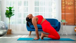 Woman on her knees on a yoga mat leaning back with her hands on the floor behind her feet. She is wearing matching red sportswear and is in a domestic setting.