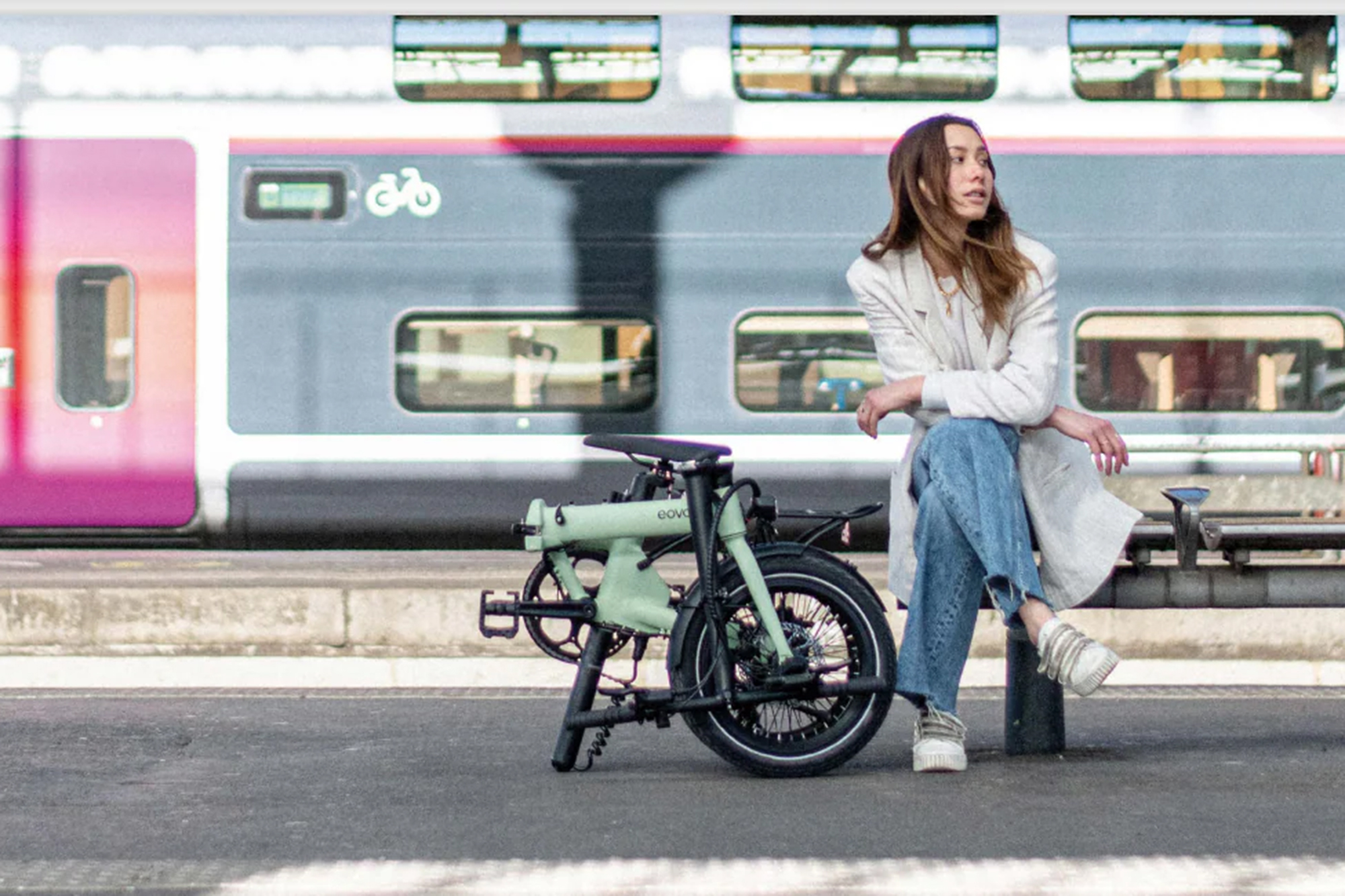 Eovolt Afternoon 16 folding bike next to a lady sitting on a bench at a train station with a double-decker train passing behind her.