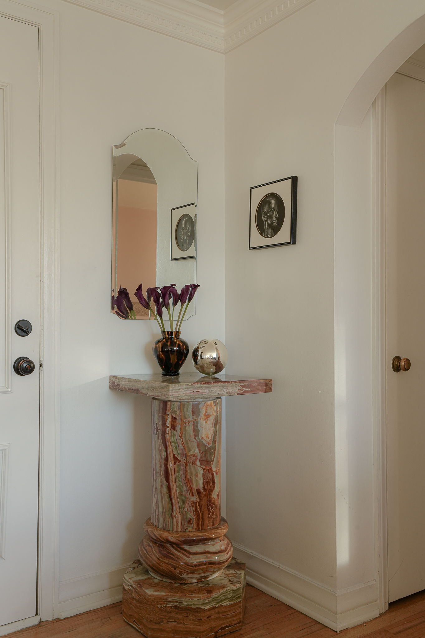 A white entryway in a small apartment. There is a Pink marble side table with a vase on it and a mirror and picture hanging over it.