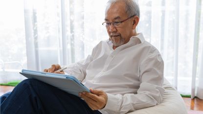An older man looks over information on a tablet while sitting in his living room.