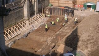 People in yellow safety vests work at a dirt covered medieval cemetery in Longwall Quad, Magdalen College under excavation.
