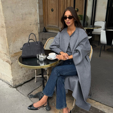 An image of a French influencer sitting at a cafe wearing a collarless coat with dark-wash jeans and black ballet flats.