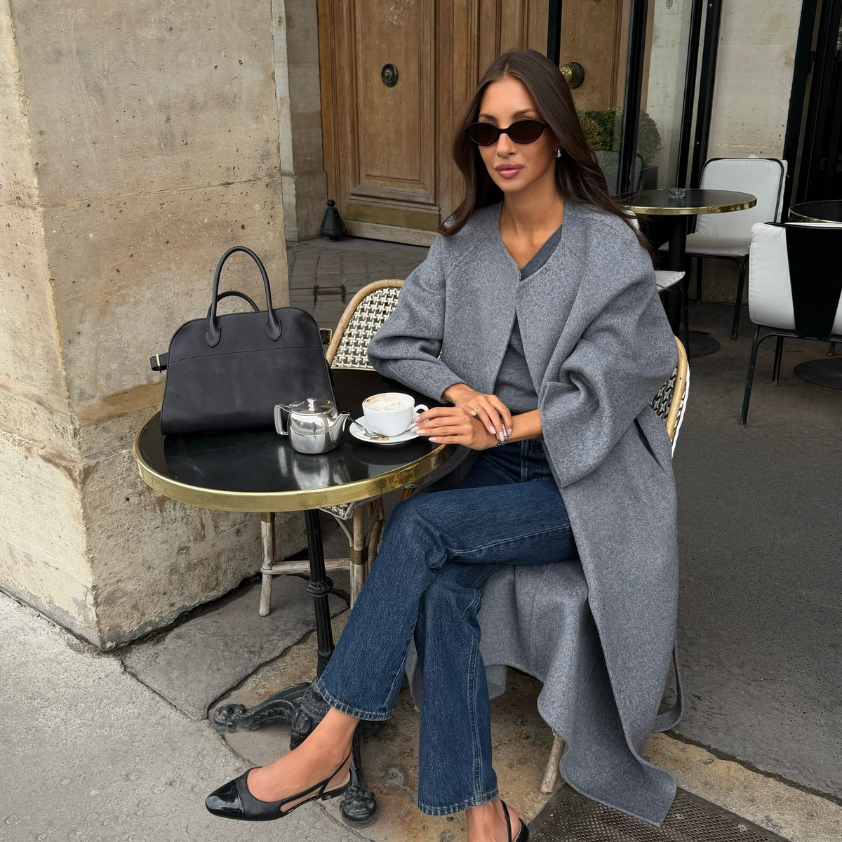 An image of a French influencer sitting at a cafe wearing a collarless coat with dark-wash jeans and black ballet flats.