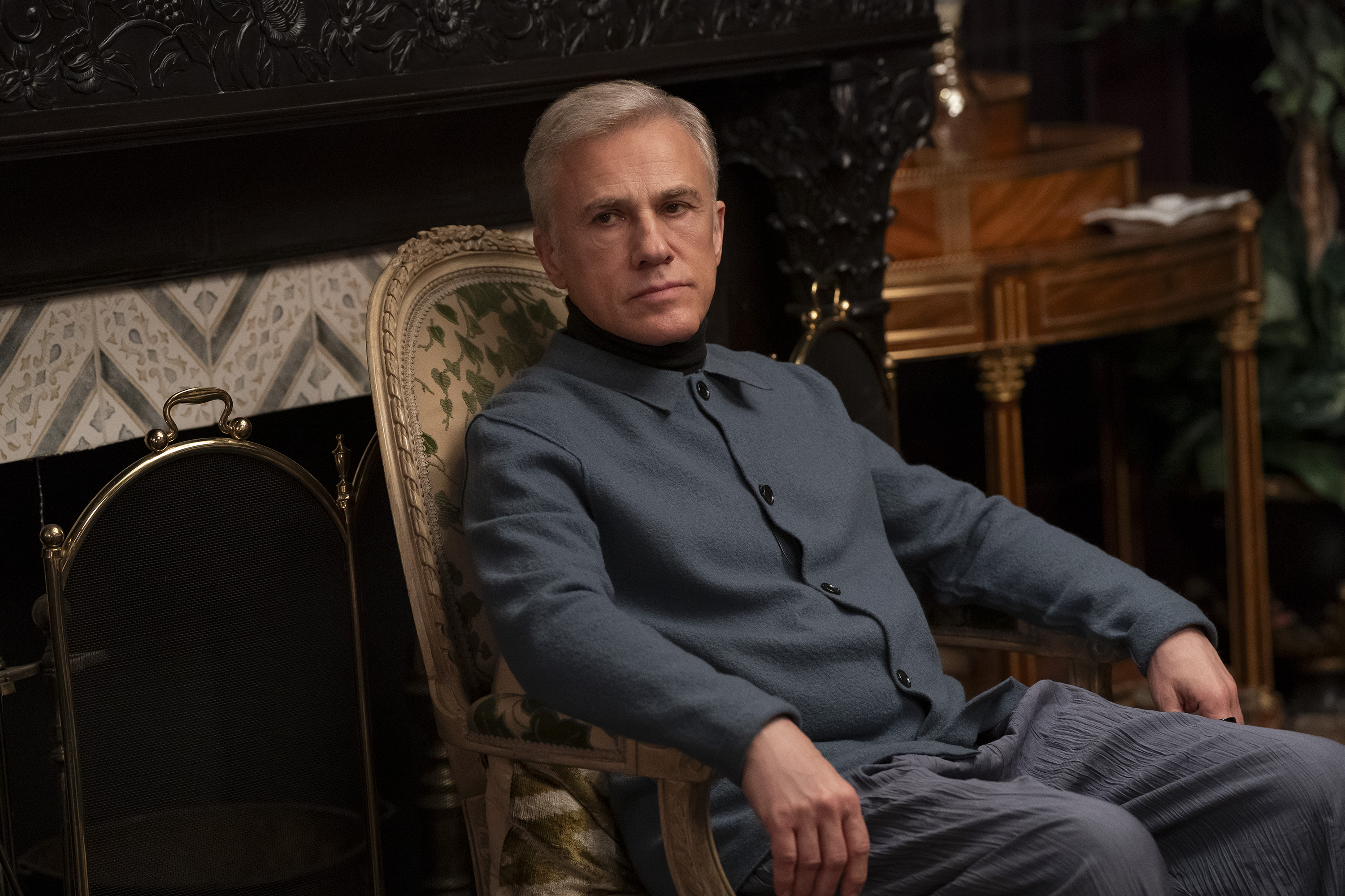 Christoph Waltz as Bash Steed, sitting in a chair in front of an unlit fireplace, in &#039;Only Murders in the Building&#039; season 5
