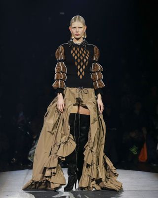 Andreas Kronthaler for Vivienne Westwood S/S 2023. A female model wearing a black patterned top with puffy brown patterned sleeves, a long brown dress and thigh high black boots.