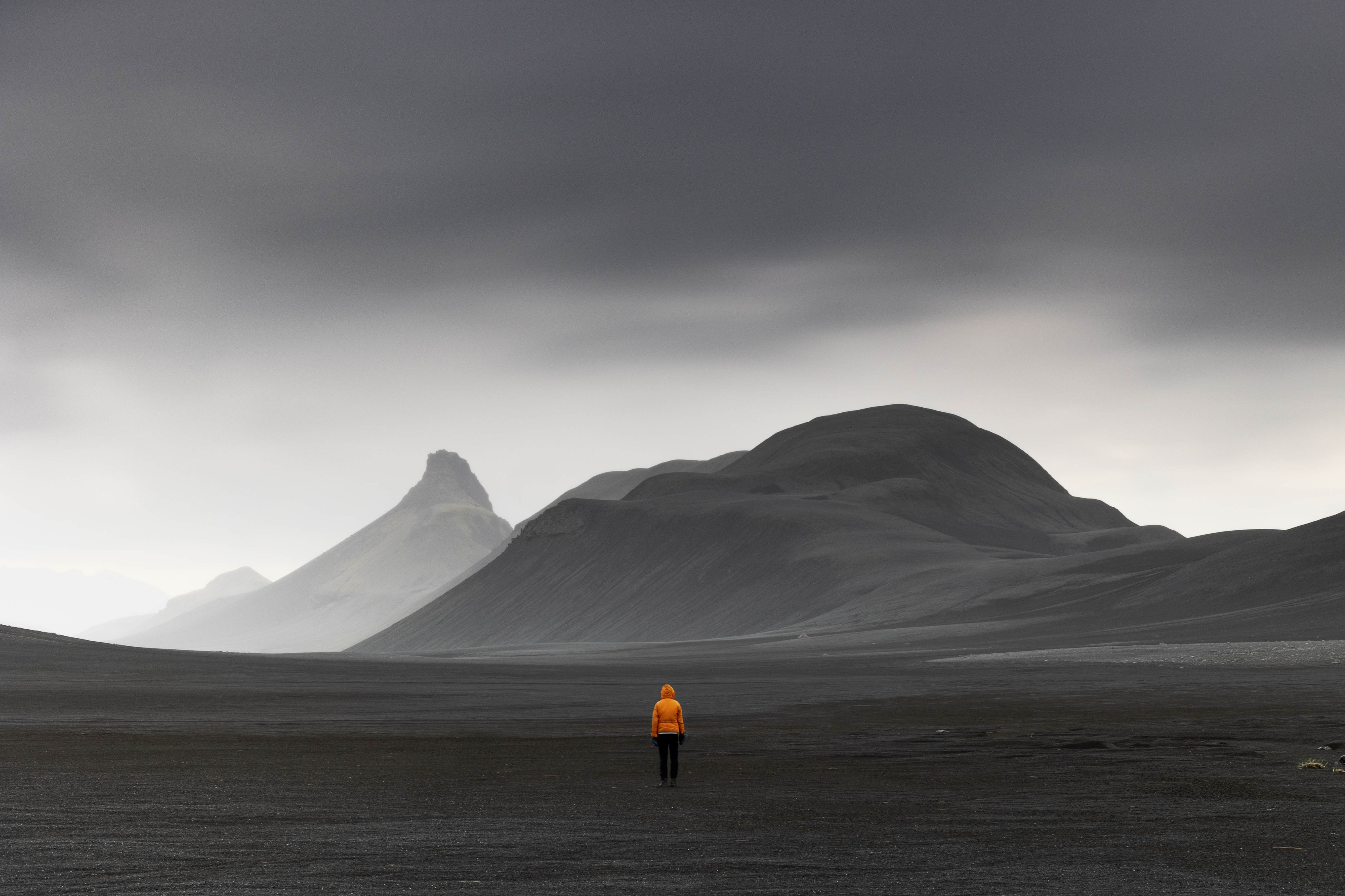 A photograph of a person dressed in a bright orange jacked against a backdrop of black landscape in Iceland