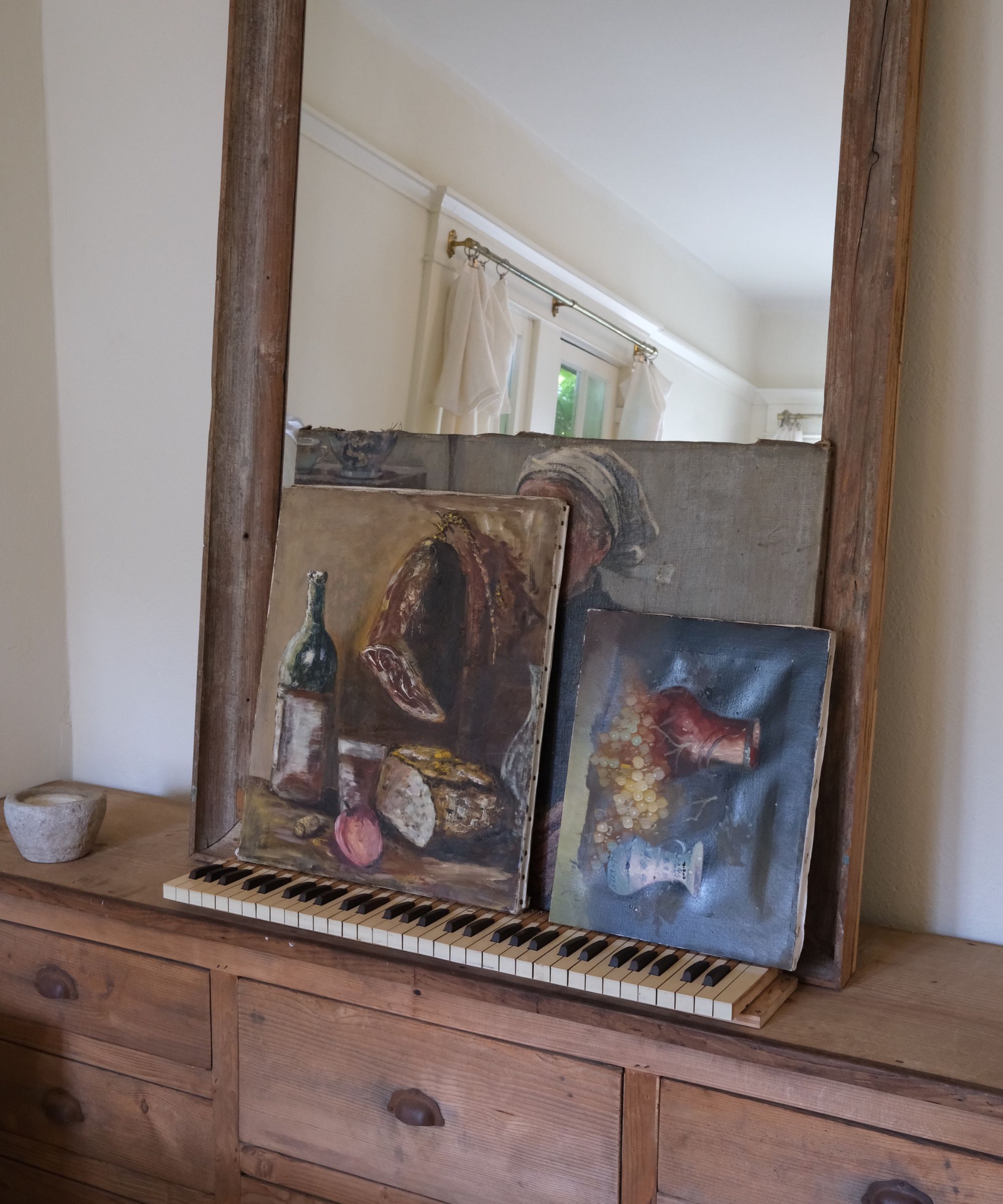 Still life oil paintings rested against a rustic mirror on a wooden dresser, supported by an antique piano keyboard base.