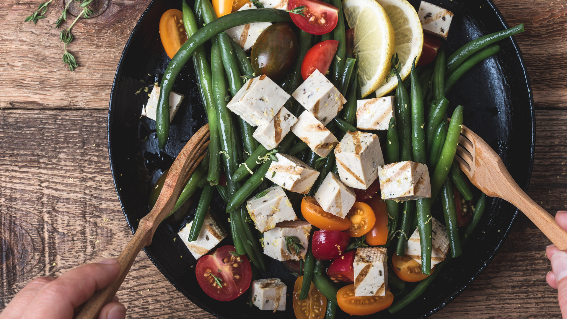 Delicious summer vegan meal, cooking healthy green beans salad with grilled tofu, fresh colorful mix cherry tomatoes, thyme herbs and lemon zest served in rural cast iron skillet, wooden forks, top view