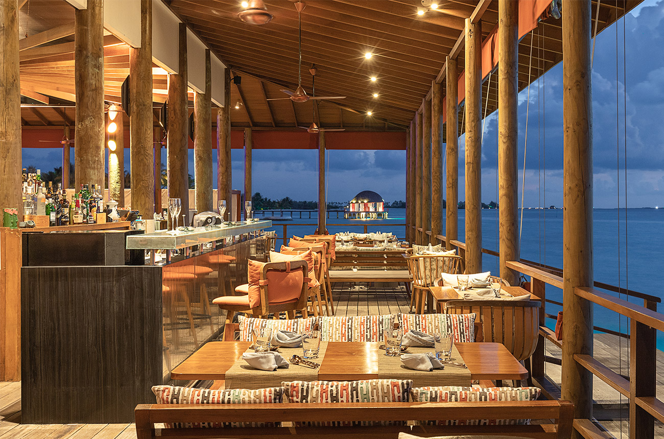 Image of a half-inside, half-outside dining area at dusk, with wooden tables and the sea in the background, and a bar to the left of the image