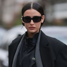 a woman with black sunglasses, dark hair with a centre parting, a black lacoste top and two tailored jackets - le labo the noir 29