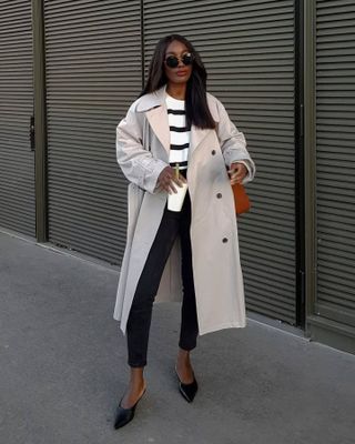 French influencer Aida Badji Sané wearing a trench coat, striped sweater, skinny black jeans, and black mule pumps