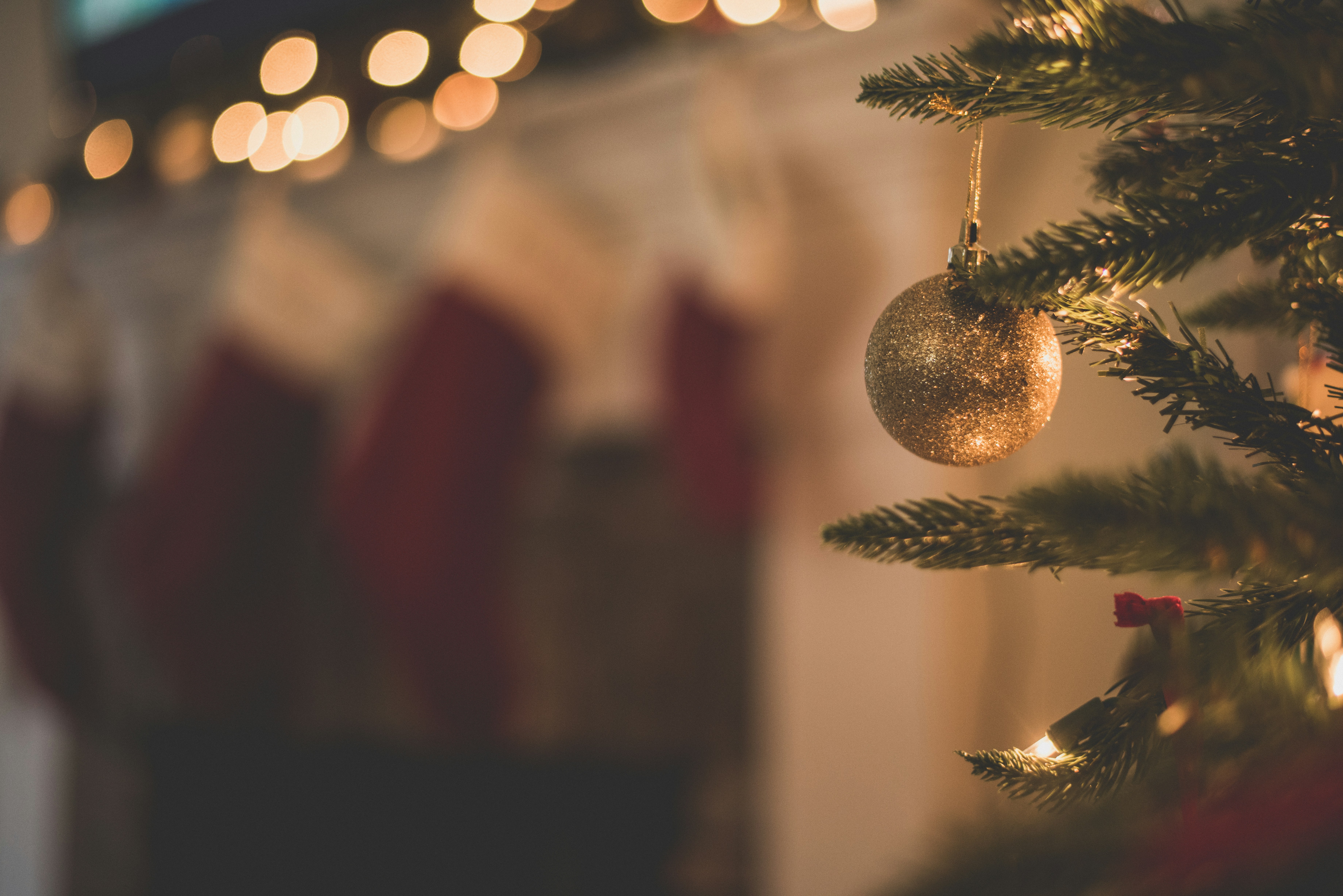 A close-up, warm-toned photograph of a sparkly gold Christmas ornament hanging on a pine tree, with out-of-focus red stockings hanging in the background and soft, warm lights blurring the top.