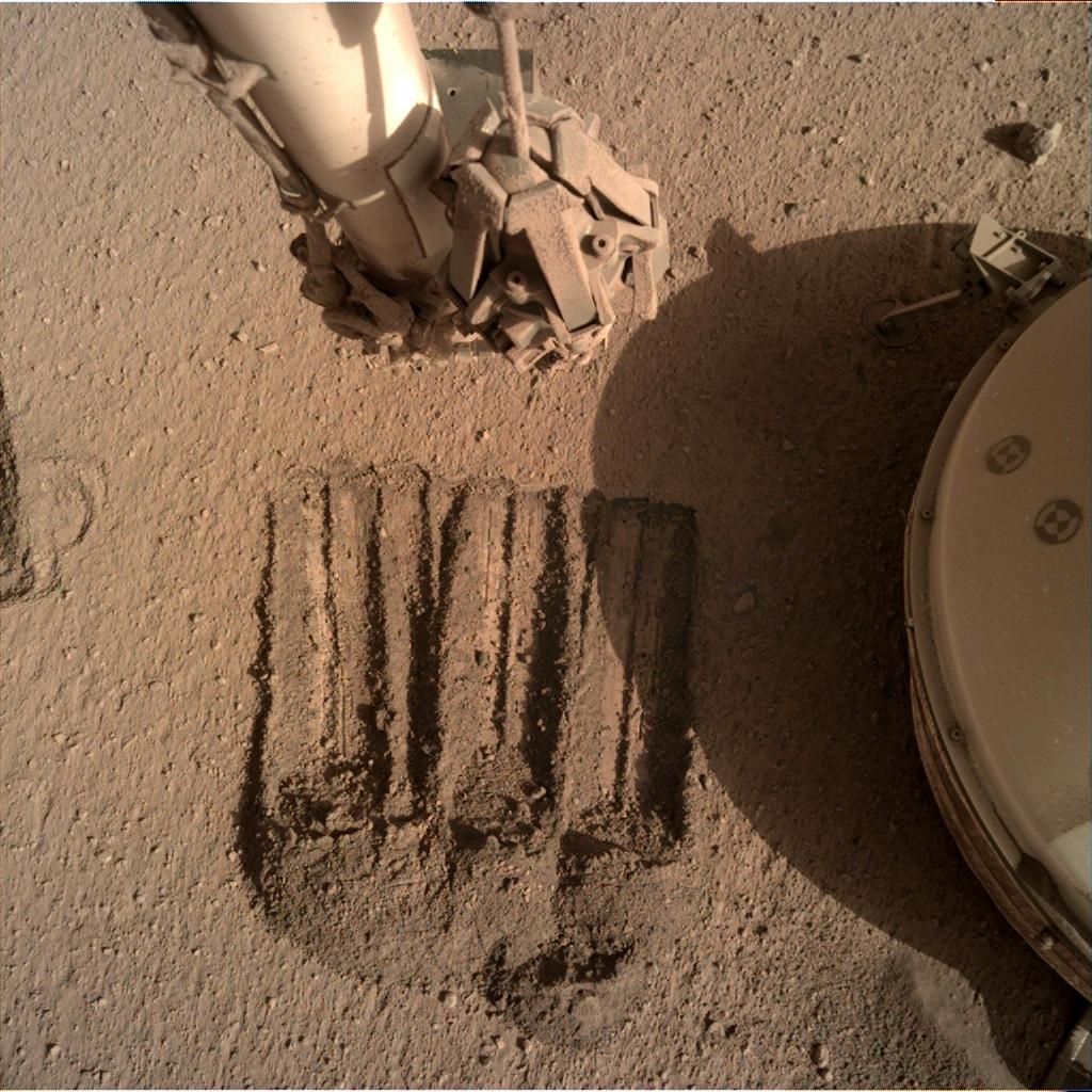 The InSight lander is using its robotic arm to scoop up Martian soil, as seen on the left, and gently cover the tether connecting the seismometer, located under the shield to the right, to the main lander. Mission personnel hope the measure will reduce noise in the seismometer&#039;s data.