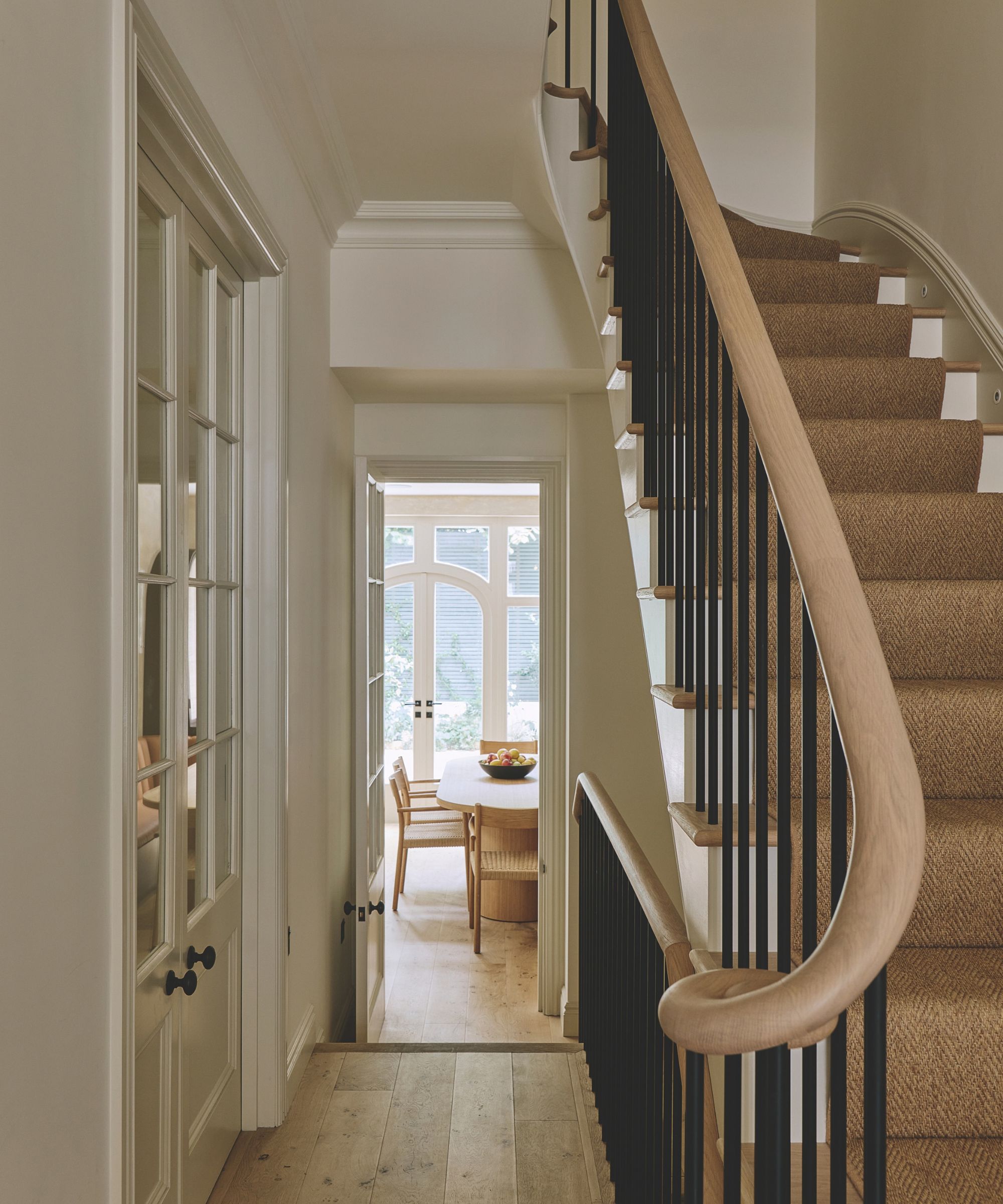 View down a home hallway featuring a light wood staircase railing with black iron spindles, sisal runner carpet, and glass double doors