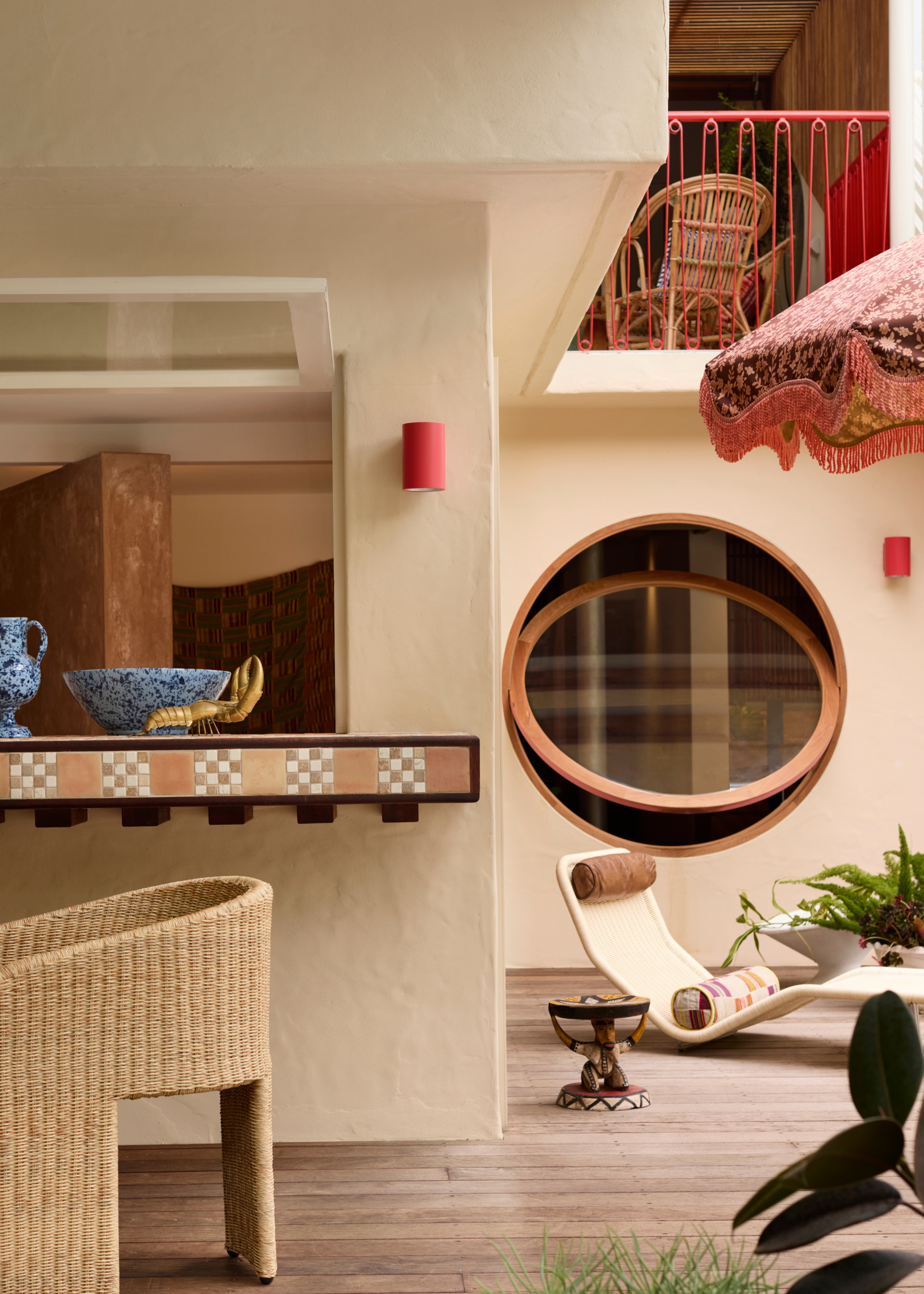An outside area with a bar ledge with a mosaic tile edging, a large porthole window half open with a sun lounger underneath, and a red-framed balustrade along the balcony area above