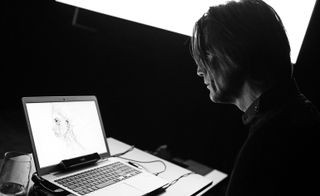 Black and white image, Fink sat at his desk looking at one of his drawings on a laptop screen, bright white projector screen in the back drop, black wall, glass of water on desk