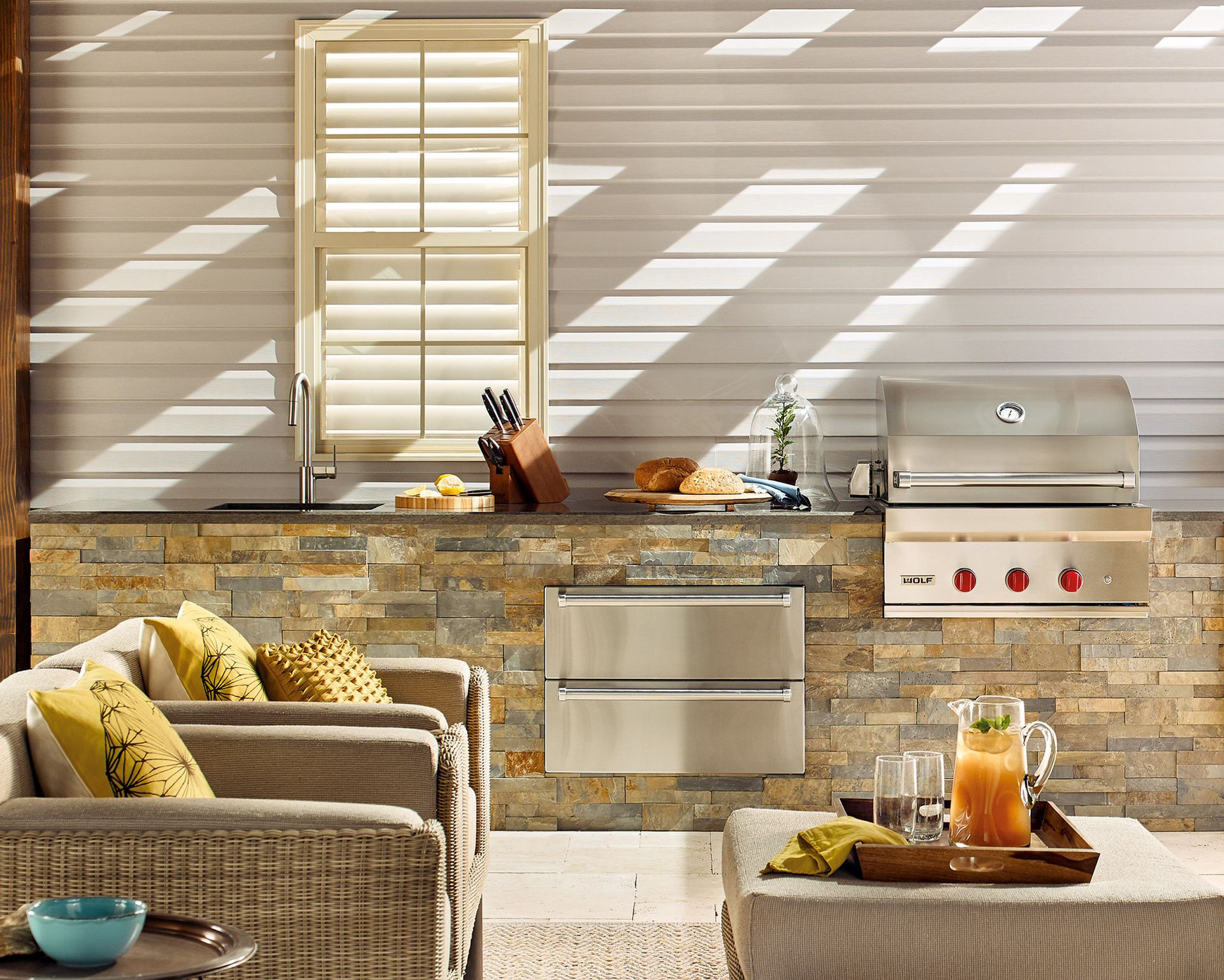 An outdoor kitchen with chrome fixtures and wicker armchair beside a white clapboard wall.