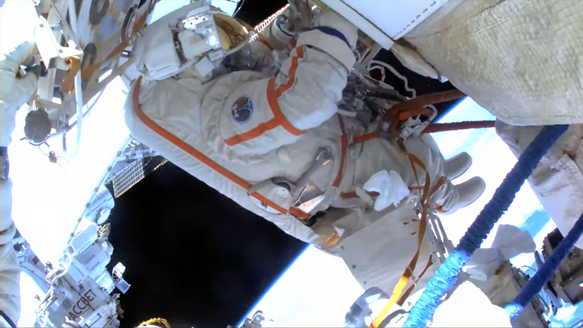 A spacesuited cosmonaut is seen from the perspective of another spacewalker's helmet-mounted camera outside a space station