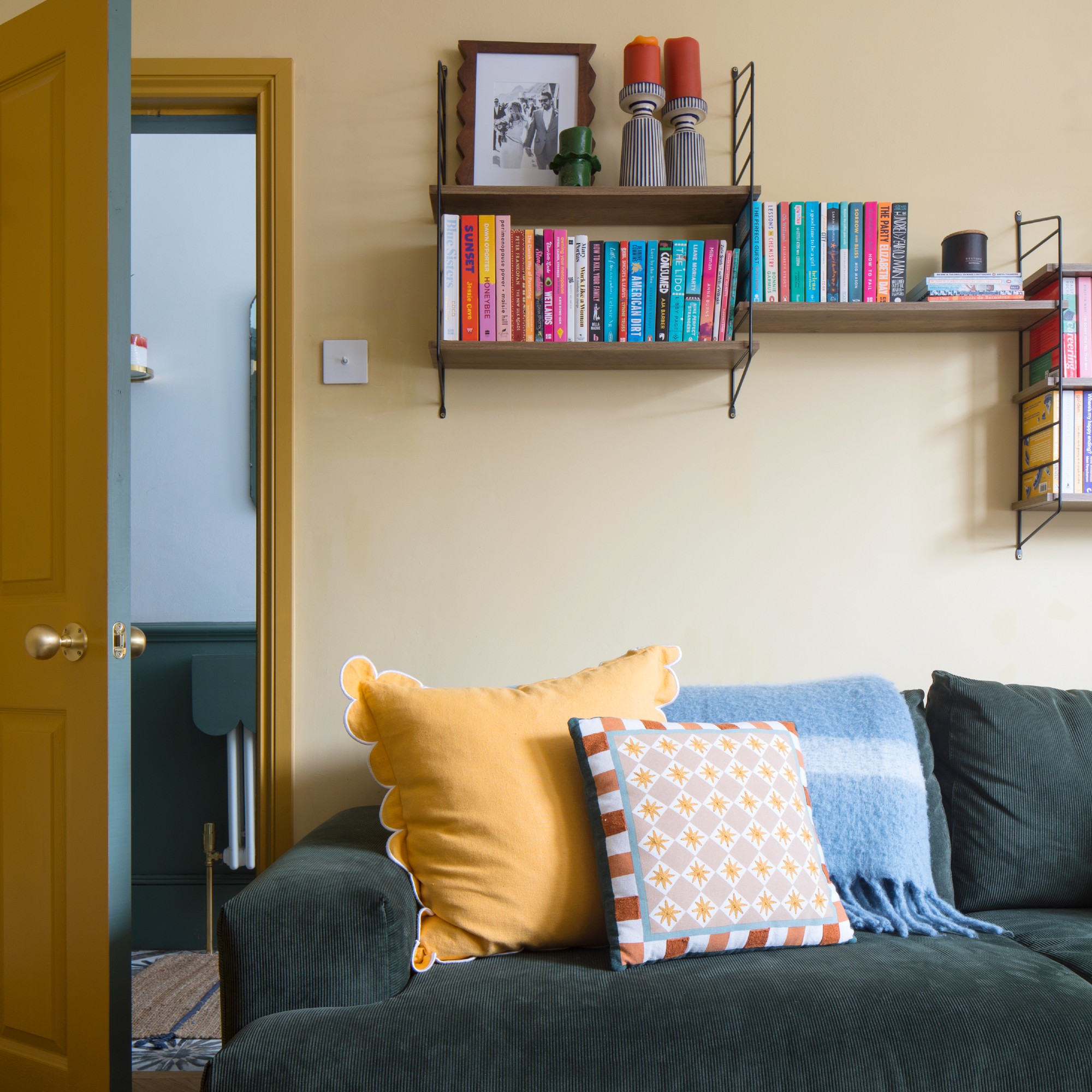 A living room painted in butter yellow with a dark corduroy sofa adorned with a yellow scatter cushion 