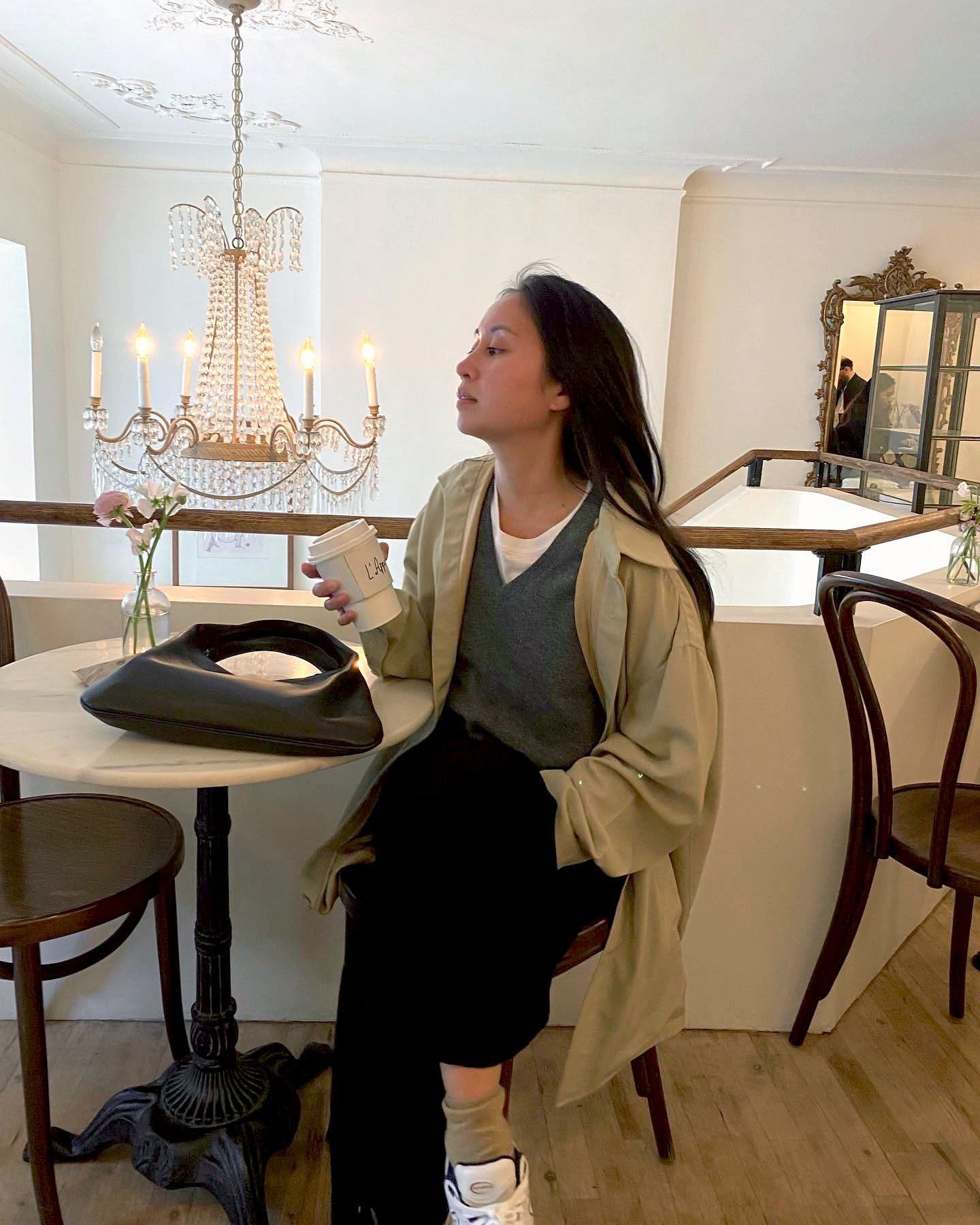 Jordan Risa Santos wearing a tan trench coat over a gray v-neck sweater, a white tee, and black trousers.