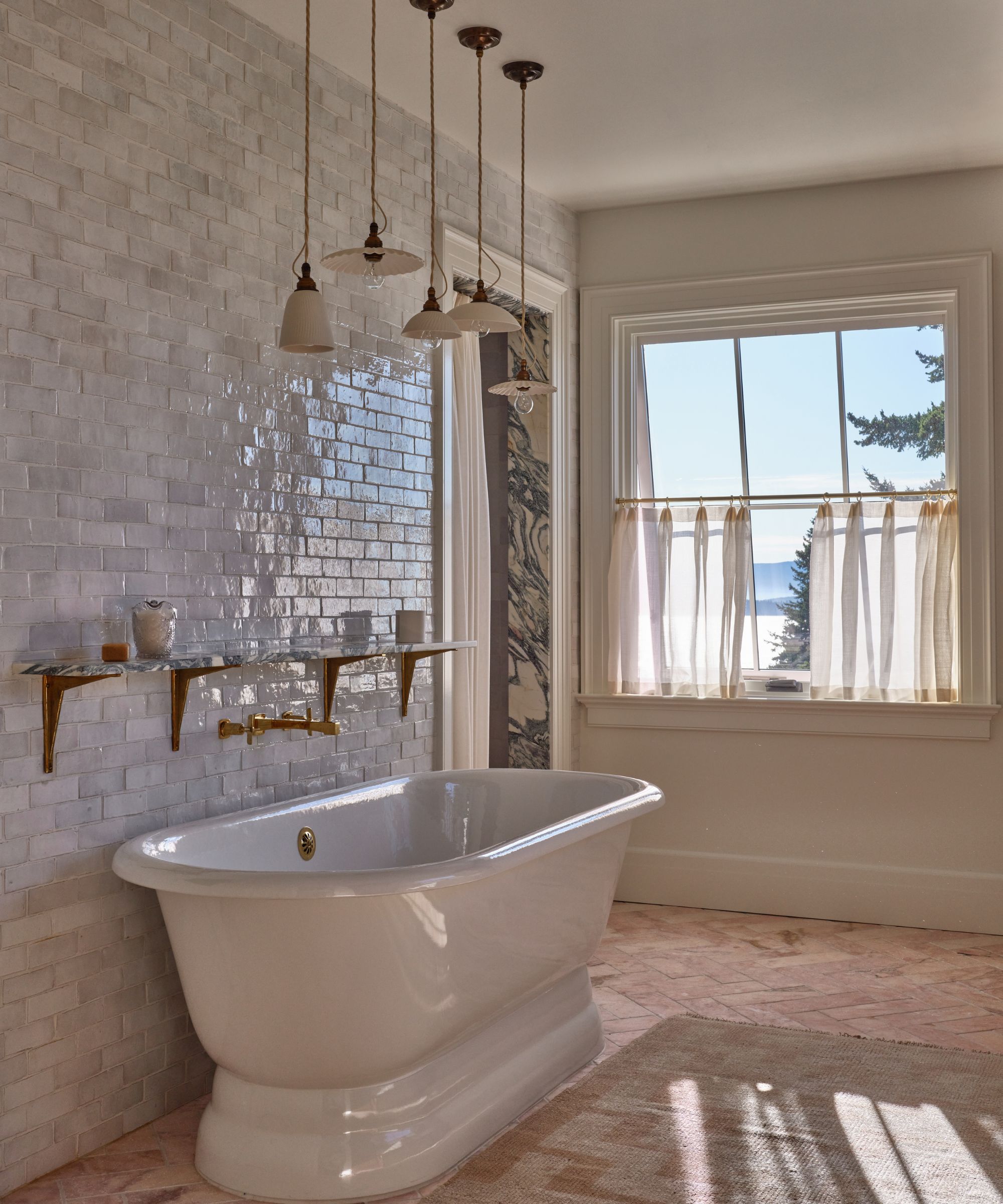 A serene bathroom centered around a white pedestal soaking tub set against a glossy off-white subway tile wall. The space includes a marble shelf with brass brackets and a cluster of delicate pendant lights