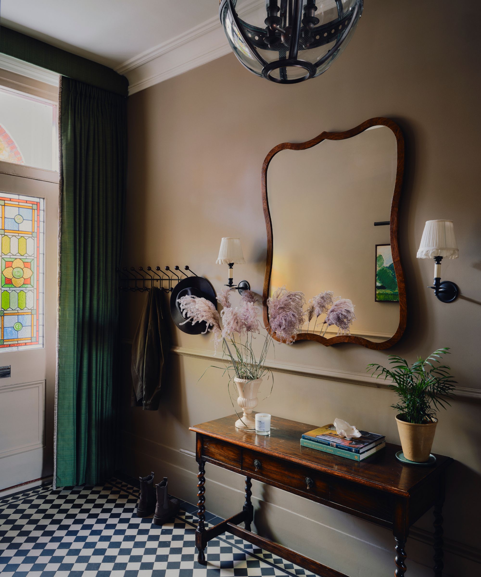 Hallway with beige walls, tiled floor, wooden console and large mirror flanked by sconces, and a stained glass front door with green drapes