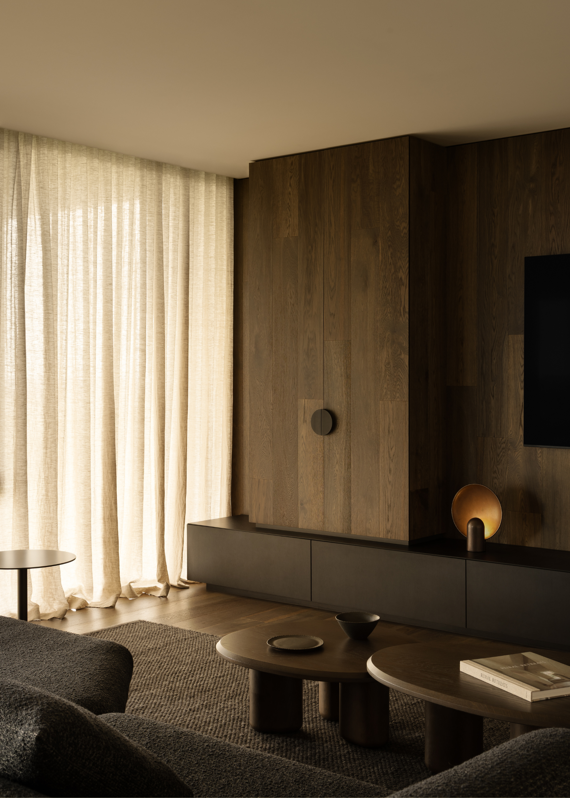 A living room corner with dark wood cupboards, a set of two dark wood coffee tables, a gray couch, and sheer curtains