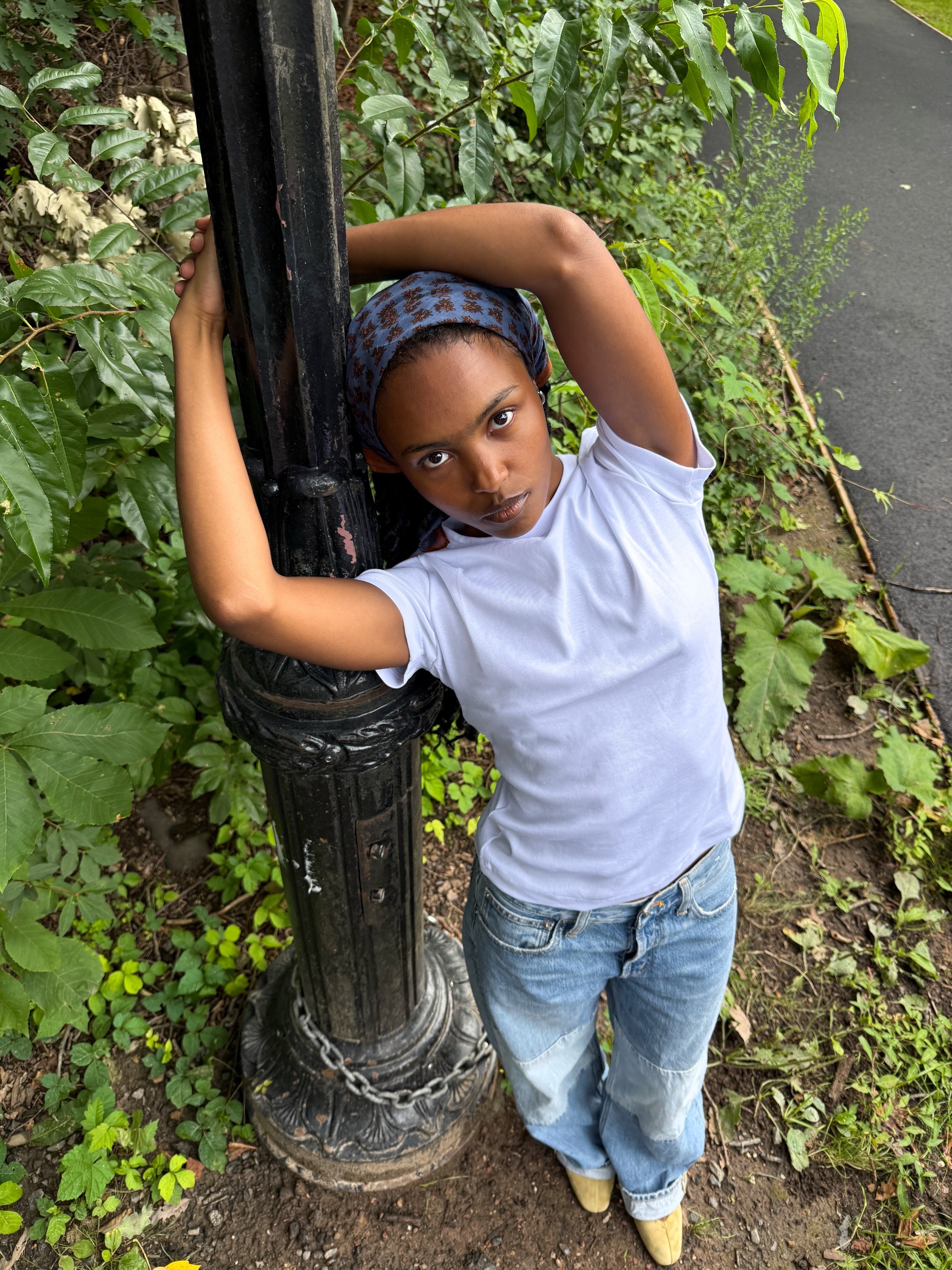 A woman wearing a bandana in her hair as she leans against a lamp pole.